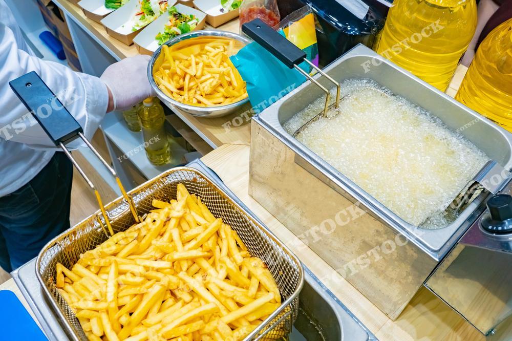 На изображении запечатлен процесс приготовления картофеля фри в коммерческой кухне. Видно, как работник кухни опускает корзину с сырыми картофельными палочками в емкость с маслом. Рядом находится ещё одна корзина с готовым картофелем фри. На столе стоят различные кухонные принадлежности и контейнеры с едой.